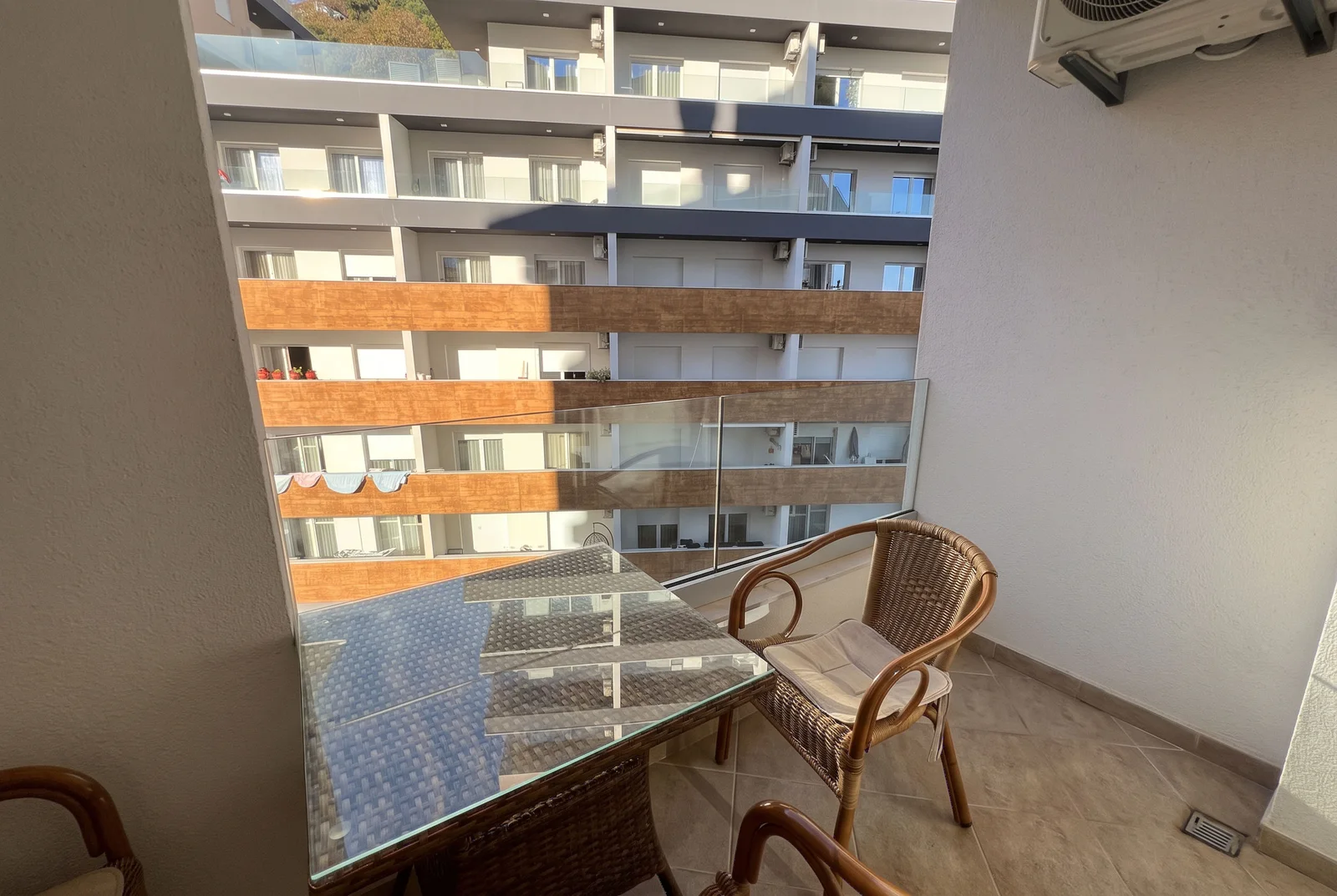 Balcony with wicker chair and glass table overlooking the facade of an adjacent apartment building.