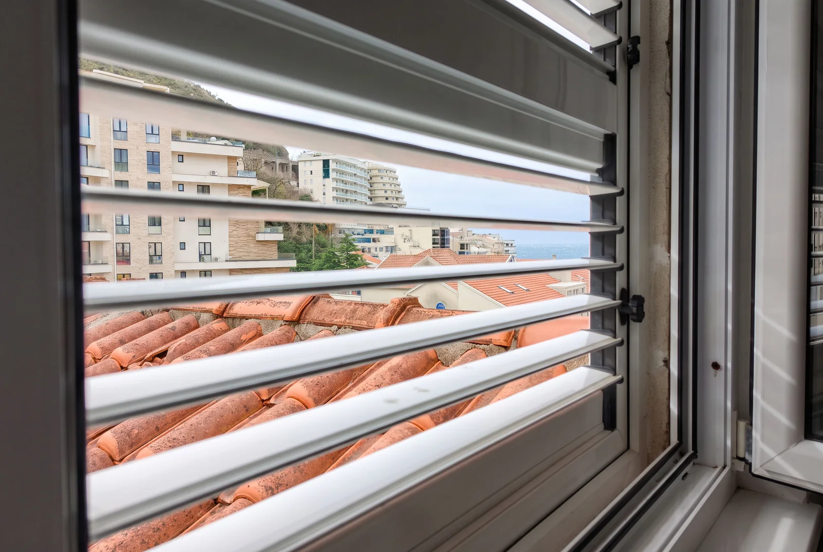 View through horizontal blinds showing a terracotta roof, city buildings, and the sea in the distance.