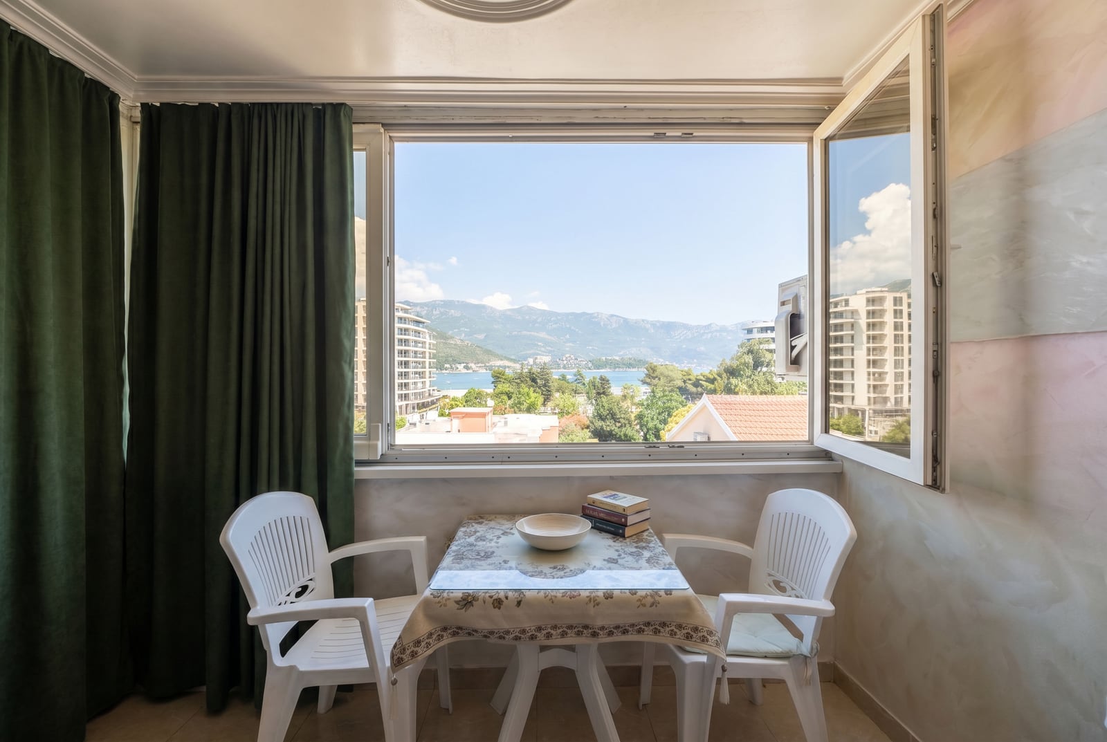 Balcony with a sea and mountain view in a Budva apartment, Montenegro: a small table with two chairs and green curtains.