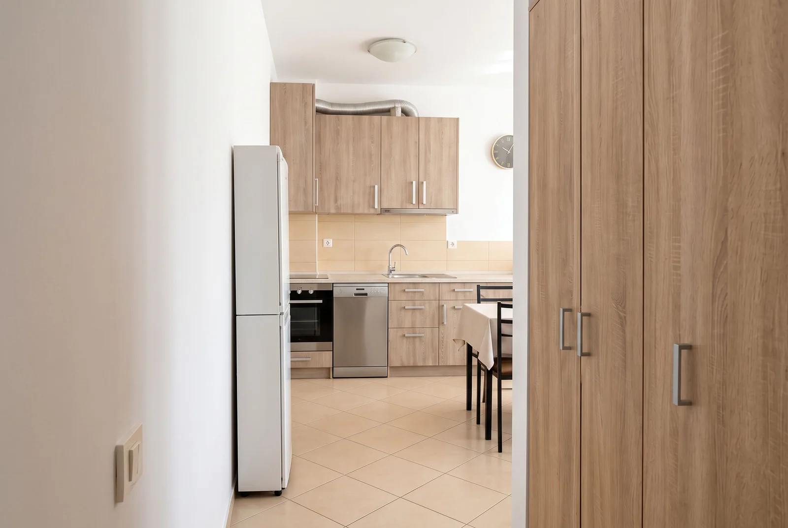 View from a hallway into a kitchen featuring light wood cabinets, a white refrigerator, dishwasher, sink, and part of a dining table.