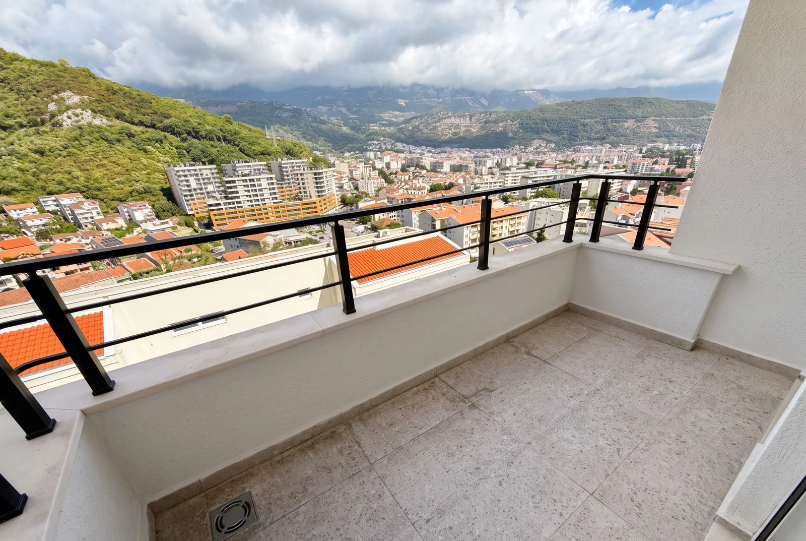 Prazan balkon sa keramičkim podom i crnim metalnim ogradama, pogled na grad i planine.