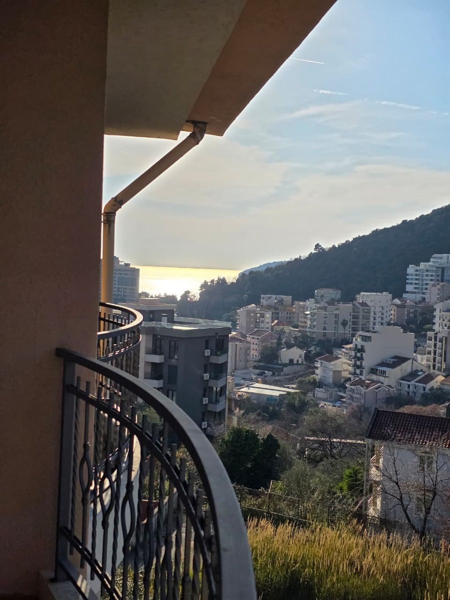 Pogled sa balkona stana u Bečićima, Crna Gora: panorama grada, planina i morske obale po sunčanom danu.