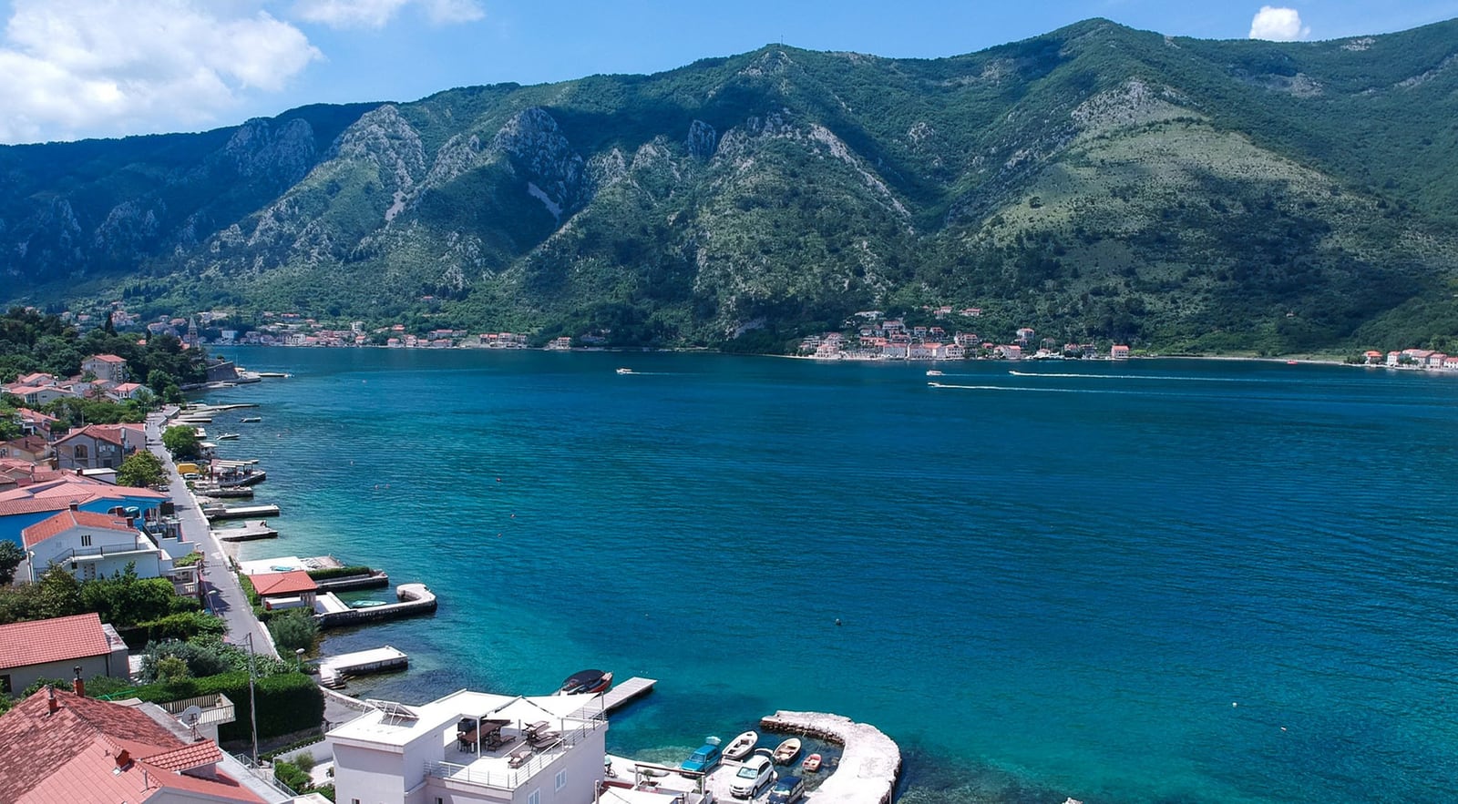 Panoramablick auf die Bucht von Kotor und eine Villa in Dobrota, Montenegro: Küstenlinie, Stege und Berge im Hintergrund.