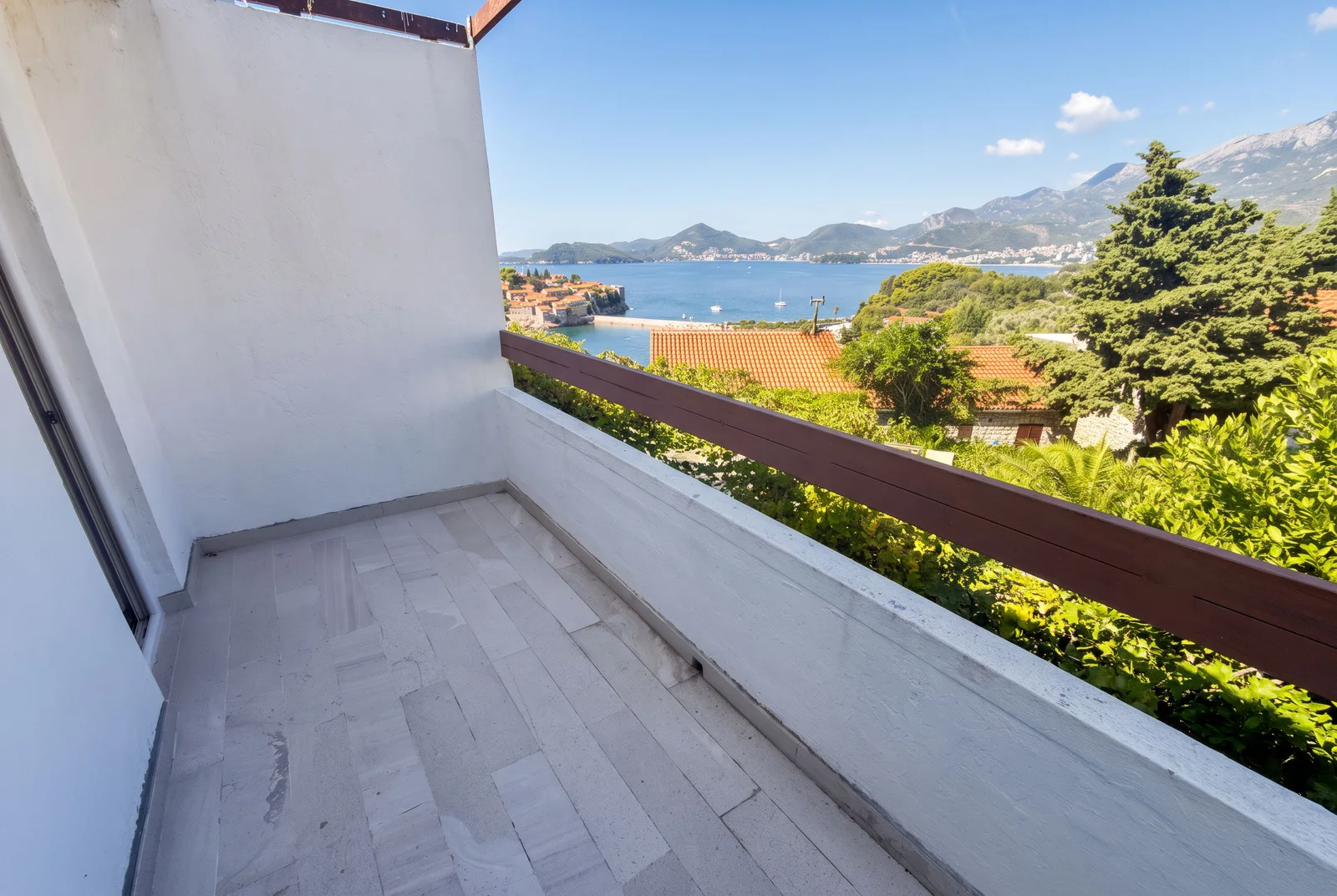 Balkon sa svetlim pločicama i drvenim ogradama sa pogledom na more, planine i historijski grad.