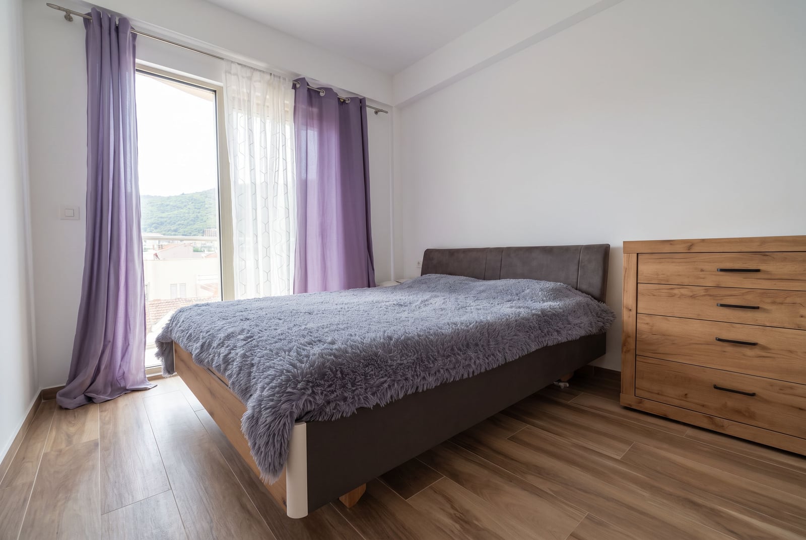 Svetla spavaća soba u stanu u Budvi, Crna Gora: bračni krevet, drveni komoda i izlaz na balkon sa pogledom na planine.