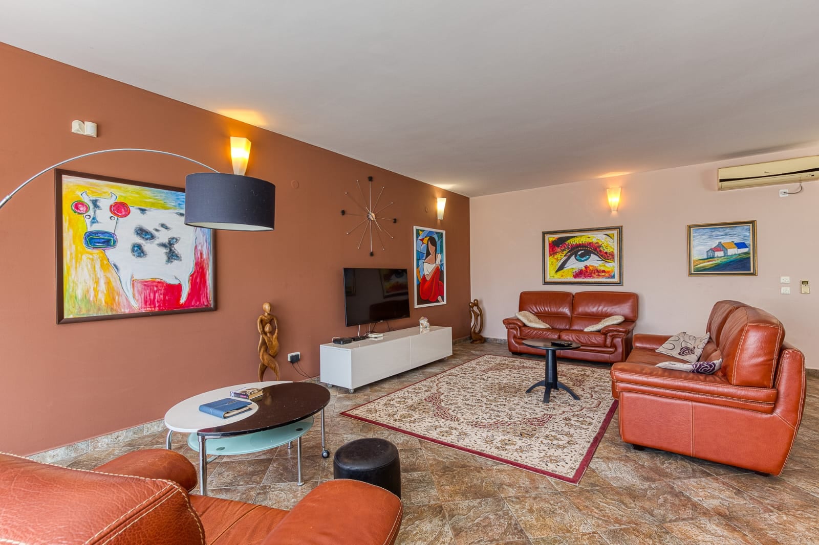 Salon avec mobilier en cuir et tableaux dans une villa à Bečići, Monténégro. Intérieur confortable avec télévision et tapis dans des tons clairs.