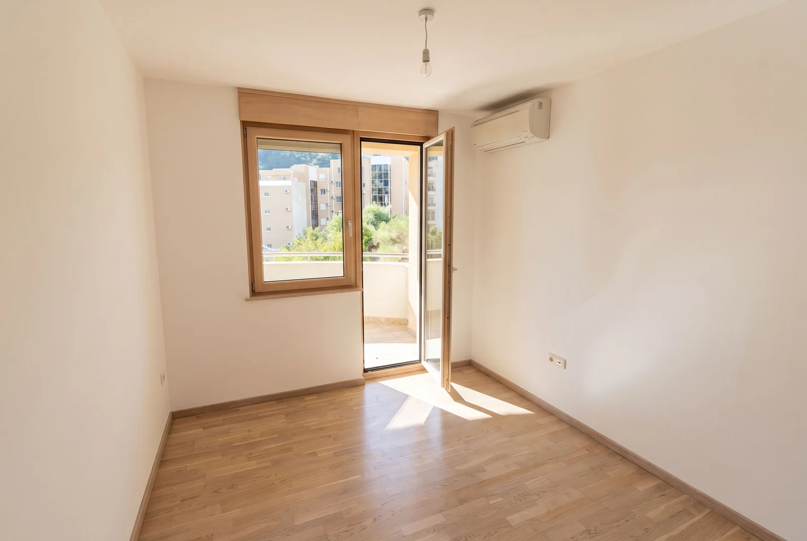 Pusta soba sa laminatom, klima uređajem i vratima na balkon. Vidljivi beli zidovi, drveni okviri i svetiljka.