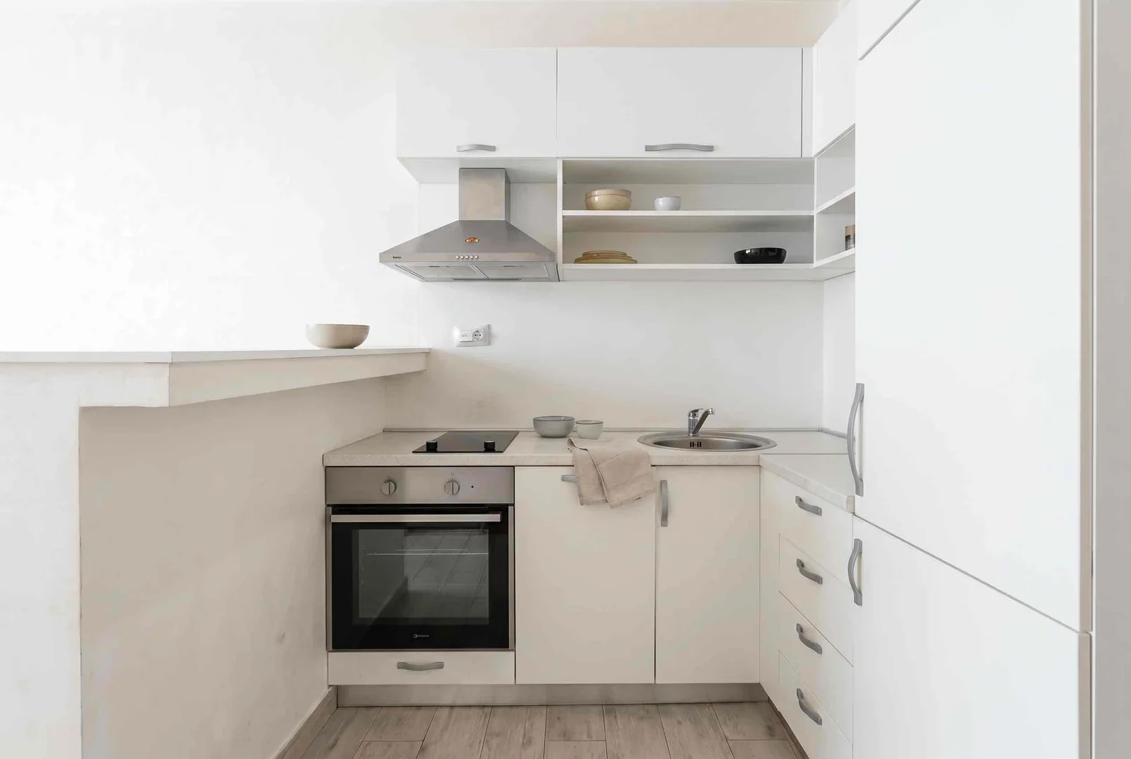 Minimalist white kitchen featuring a built-in oven, cooktop, sink, and stainless steel range hood.