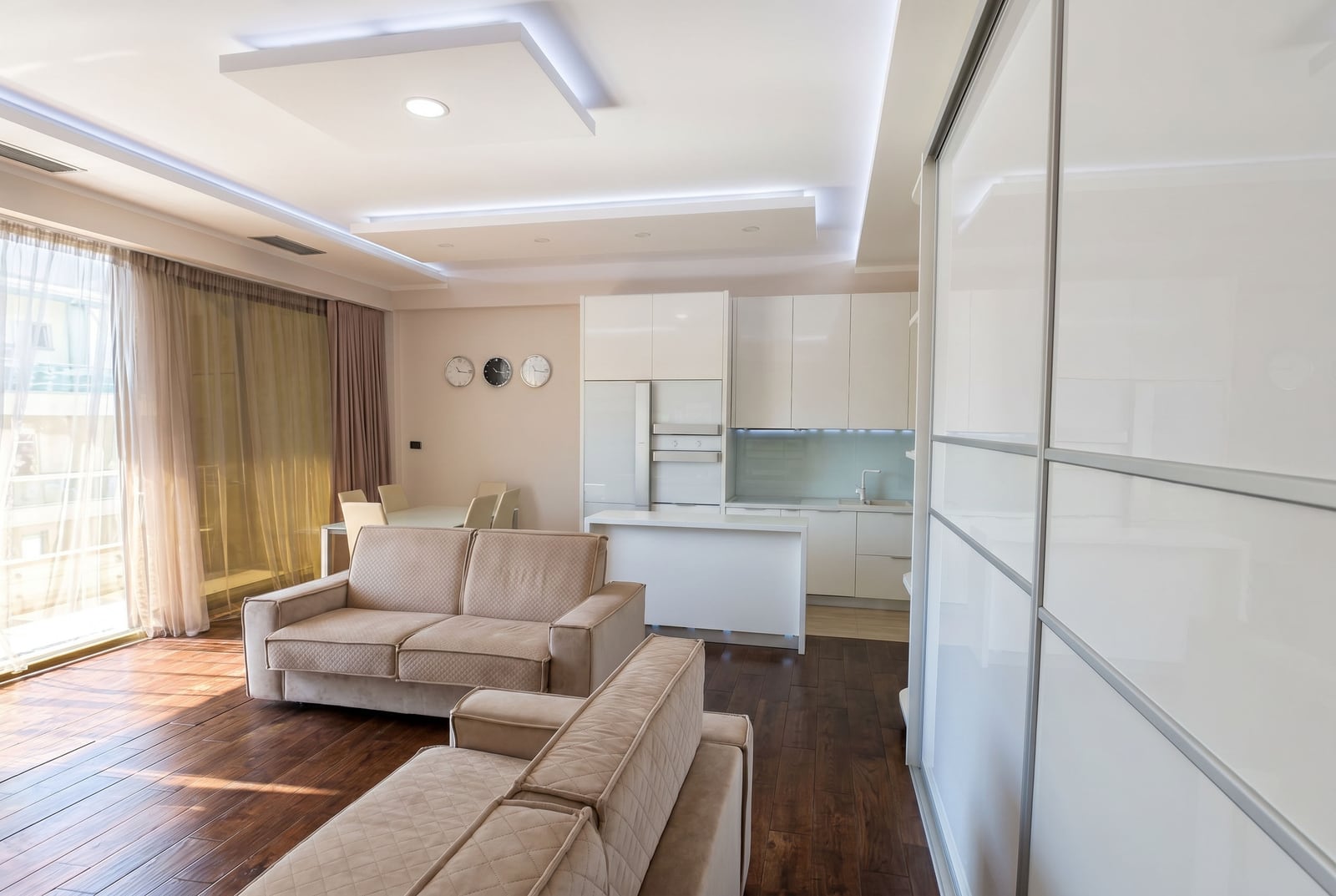 Modern living room with an open-plan kitchen in a Budva apartment, Montenegro: white furniture, light-colored sofa, and floor-to-ceiling windows.