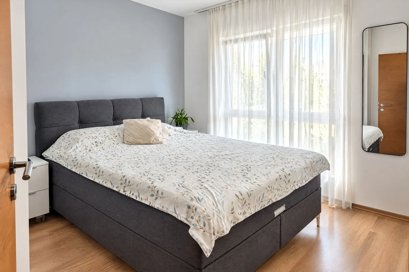 Gri kumaş başlıklı büyük bir yatak, çiçek desenli nevresim ve beyaz perdeli modern bir uyku odası.