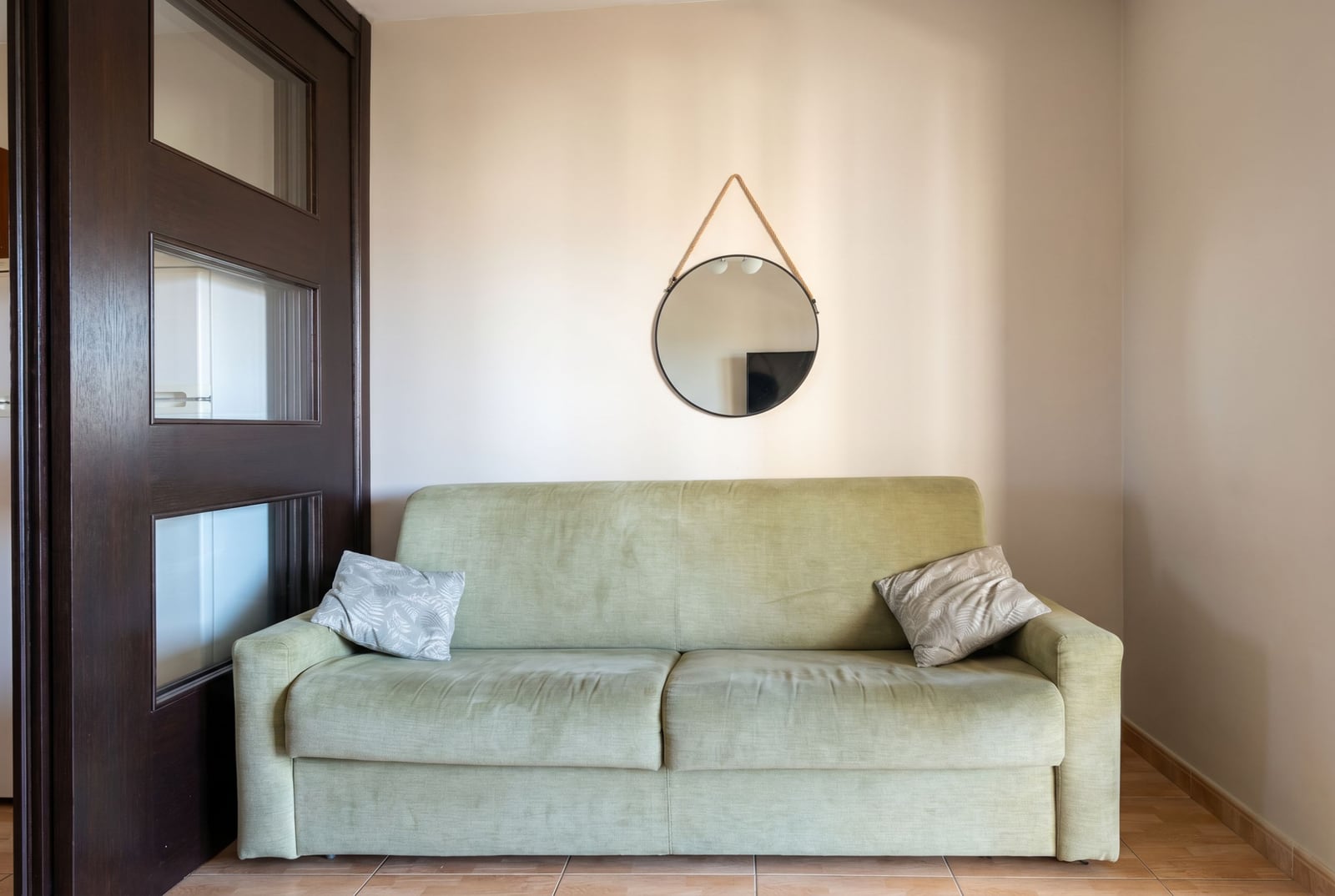 Light green sofa in the living room of an apartment in Budva, Montenegro, with a round mirror on the wall and a dark door.