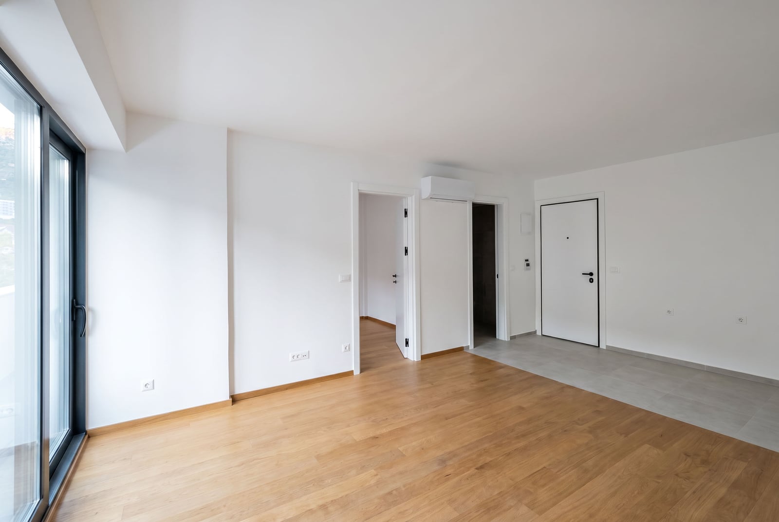 Bright empty living room with parquet flooring, panoramic window, and entrance door in a new apartment in Budva, Montenegro.