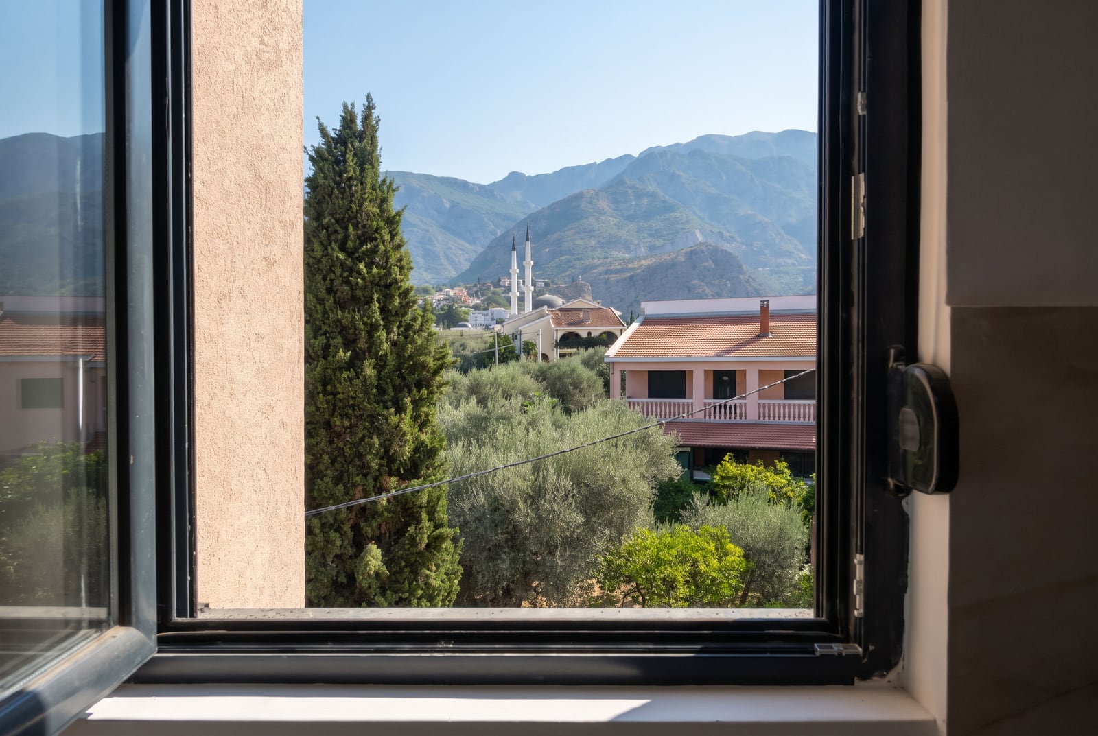 View from the bedroom window of an apartment in the city of Bar, Montenegro: mountains, olive trees, and a mosque on a sunny day.
