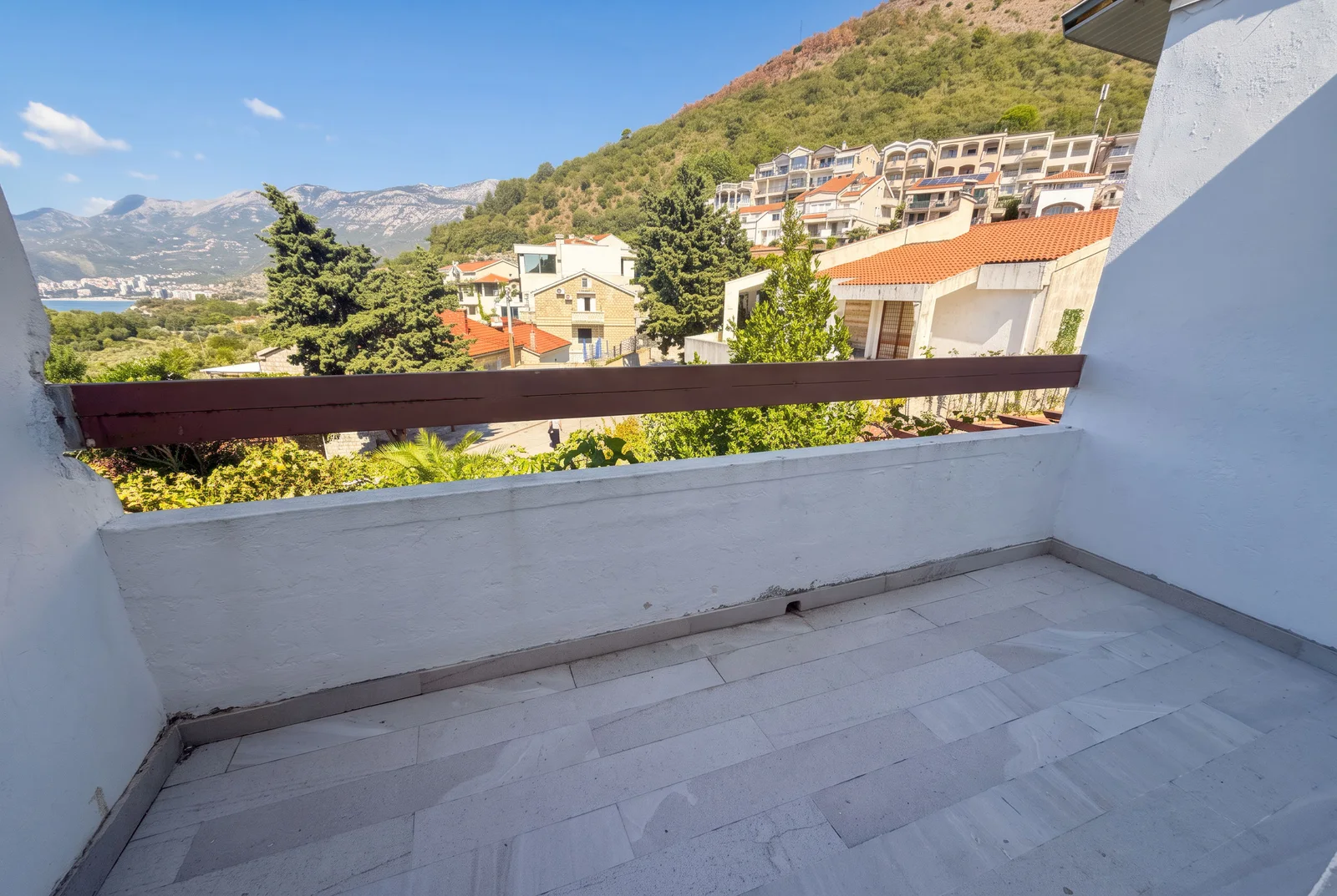 Prazan balkon sa sivim pločama i drvenom ogradom sa pogledom na planine, domove i more.
