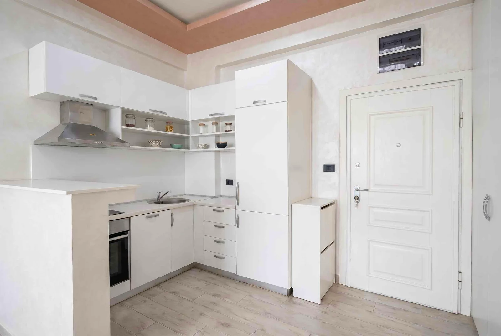 White kitchen with upper cabinets, hood, sink, oven, and a white entry door.