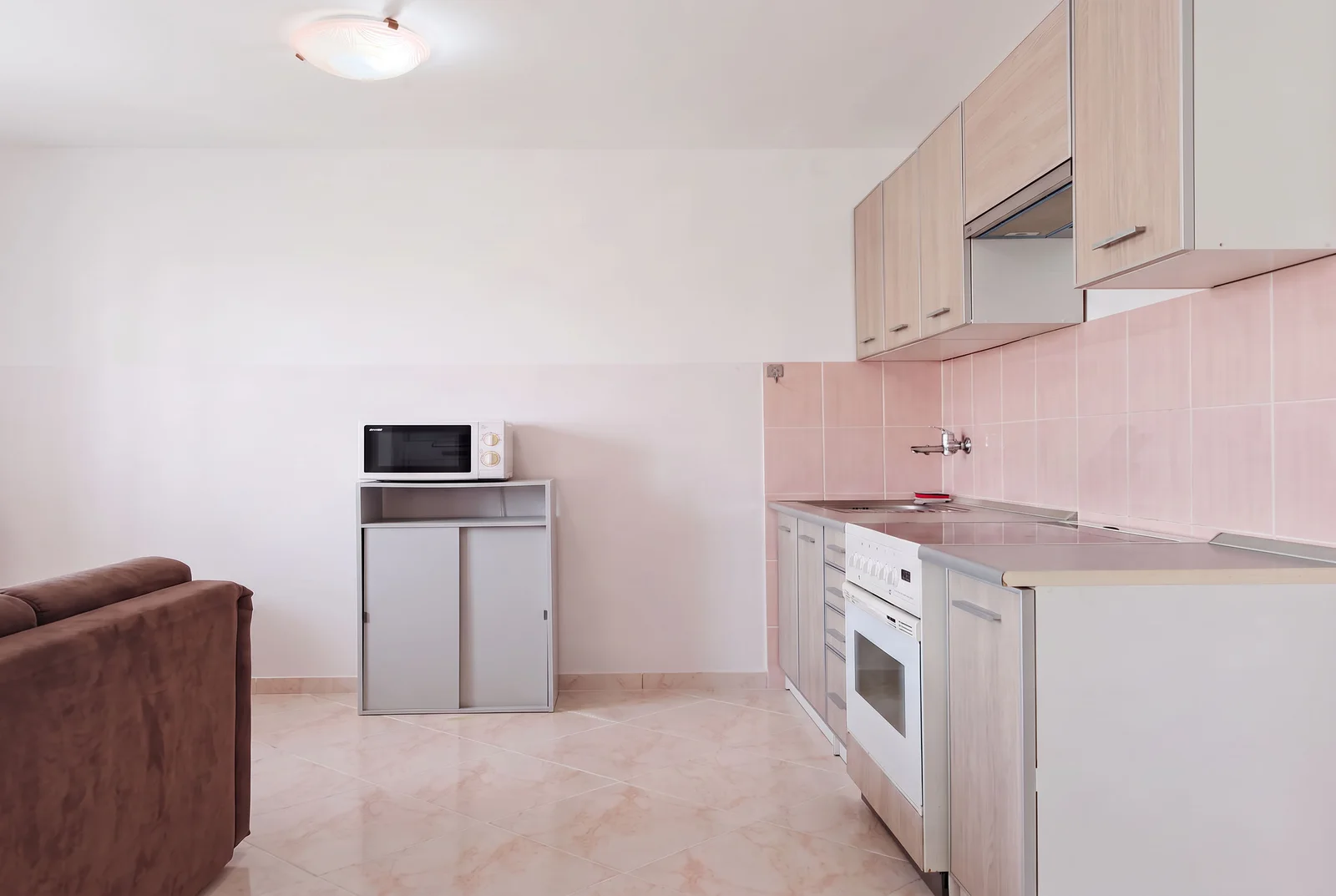 Kitchen area with light wood cabinets, white stove, microwave on a stand, and pink tiled backsplash.