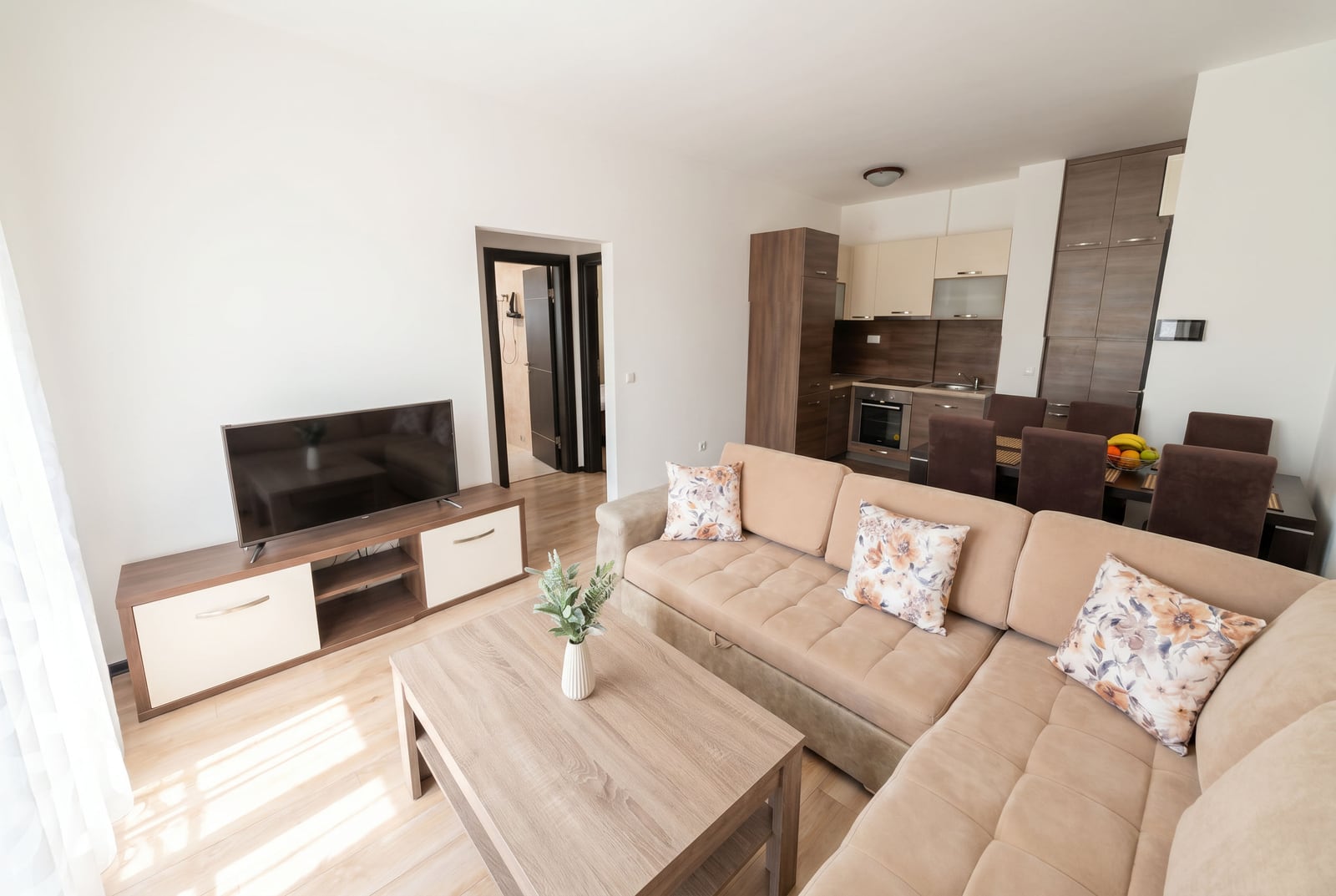 Modern living room with an open-plan kitchen in a Budva apartment, Montenegro: beige sectional sofa, dining area, and TV stand.