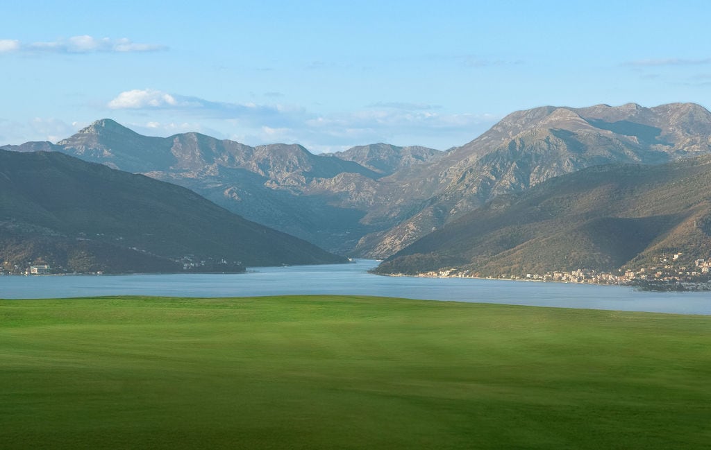Panoramic view of the Bay of Kotor and mountains from a residential complex in Luštica, Montenegro, with a green field in the foreground.