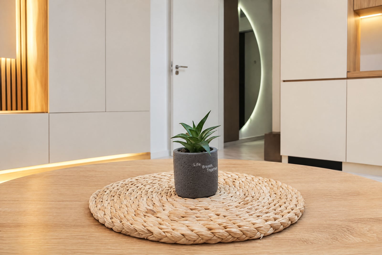 Wooden table with woven mat and small potted plant against white cabinetry and modern interior design.