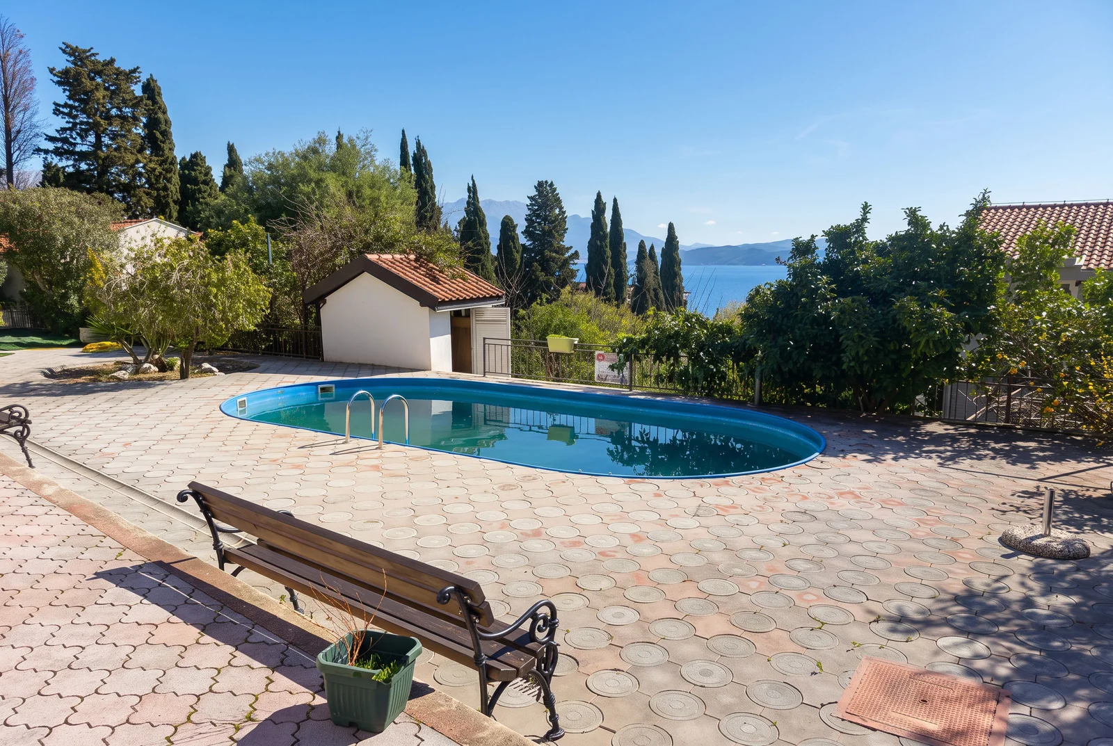 Piscine extérieure sur terrasse pavée avec banc en bois, entourée d'arbres et vue sur la mer.