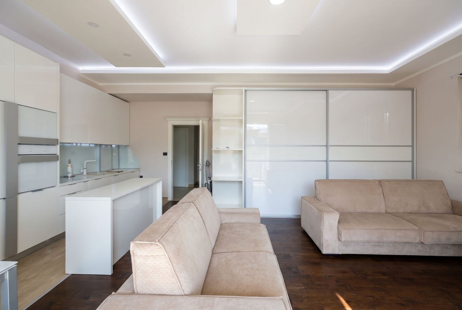 Modern living room with a white kitchen and plush sofas in a Budva, Montenegro apartment. Interior with recessed ceiling lighting.