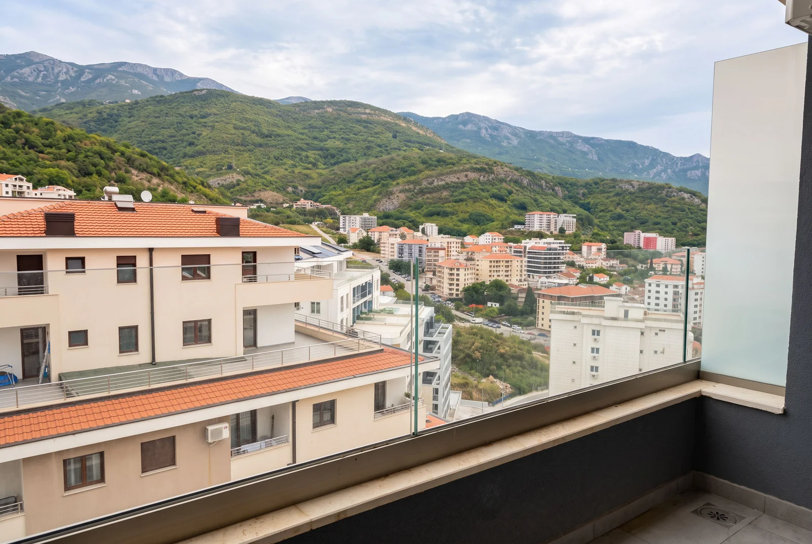 Pogled sa balkona sa staklenim ogradama na gradske zgrade i zelene planine.