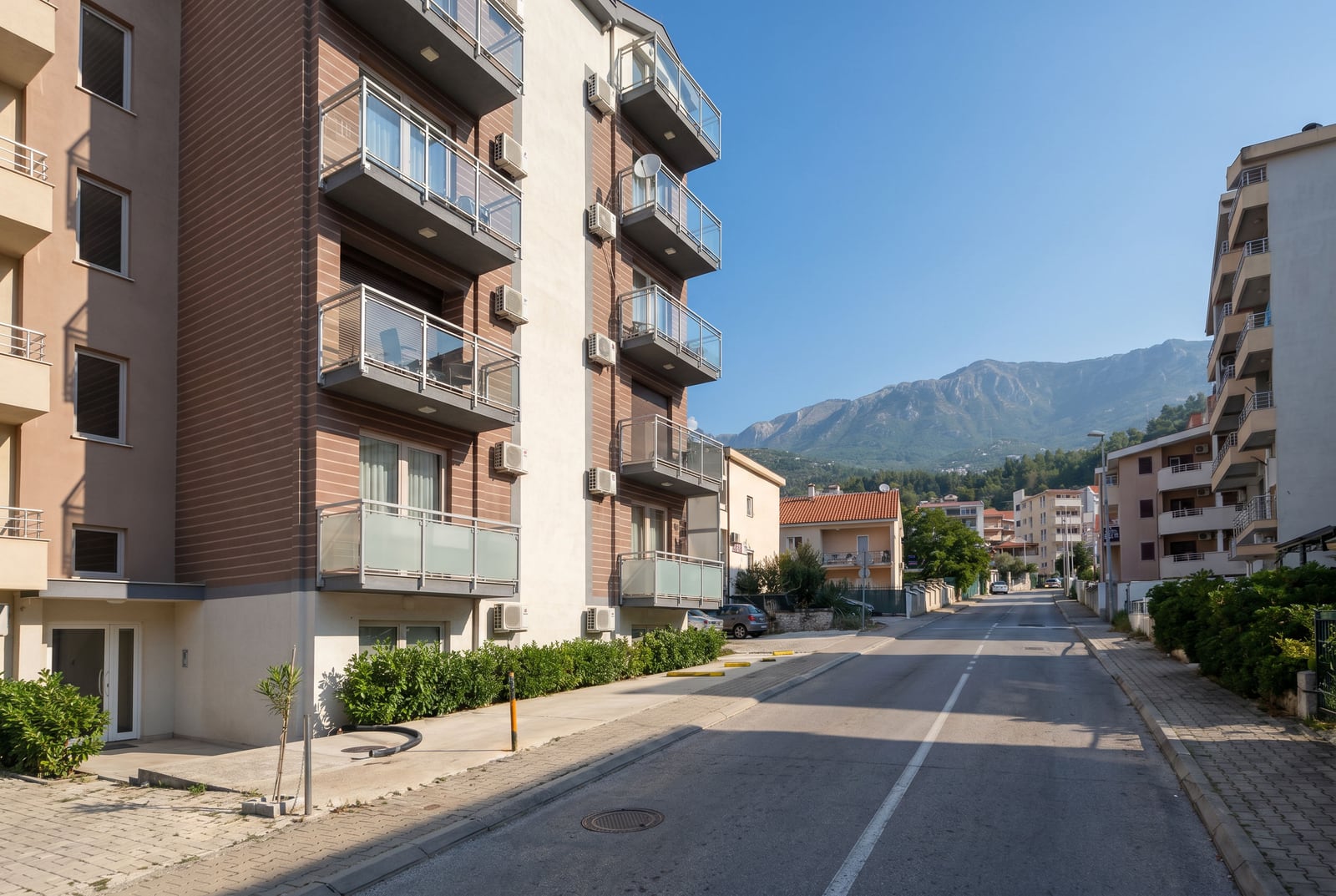 Moderna stambena zgrada sa balkonima u Budvi, Crna Gora, pogled na ulicu i planinski pejzaž u sunčanom danu.