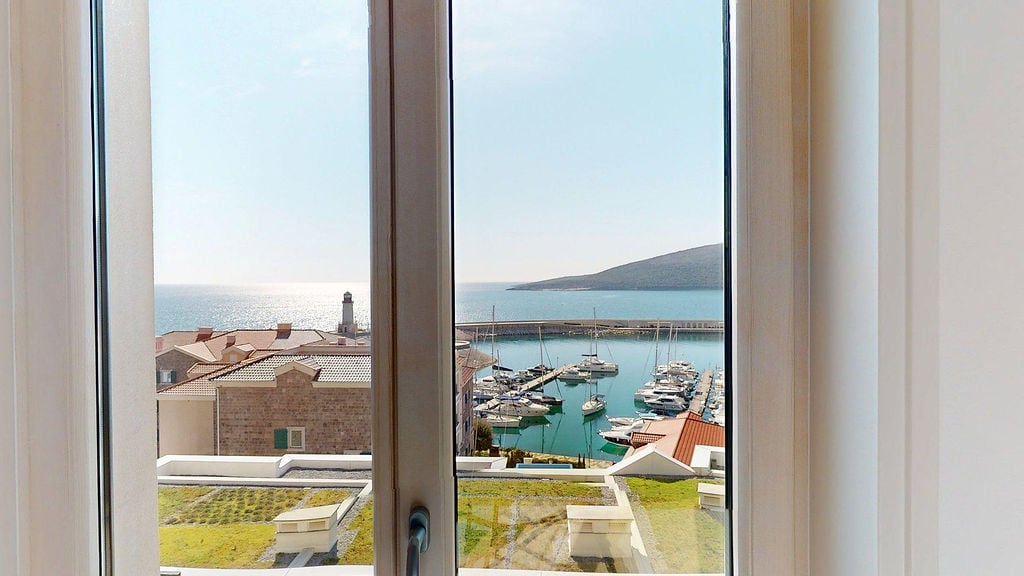 Pogled sa prozora stana u Luštici, Crna Gora: panorama na jahting marinu, more i svetionik u stambenom kompleksu.
