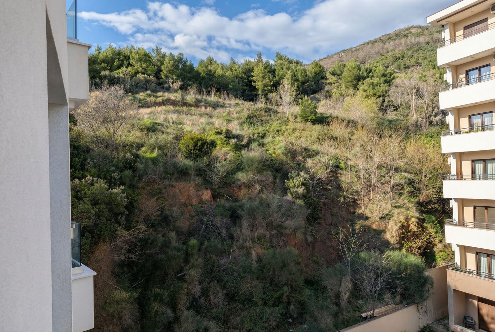 Pogled sa balkona stana u Bečićima, Crna Gora: panorama zelenog brda i susednog stambenog kompleksa sa terasama.