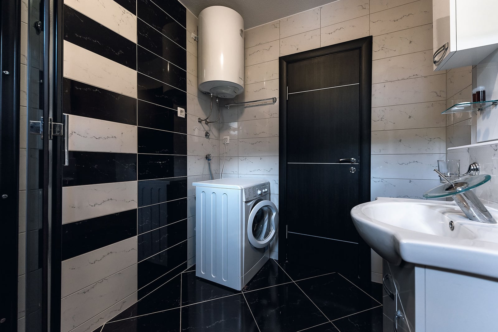 Bathroom in an apartment in Budva, Montenegro: black and white tiles, washing machine, and water heater in the interior.