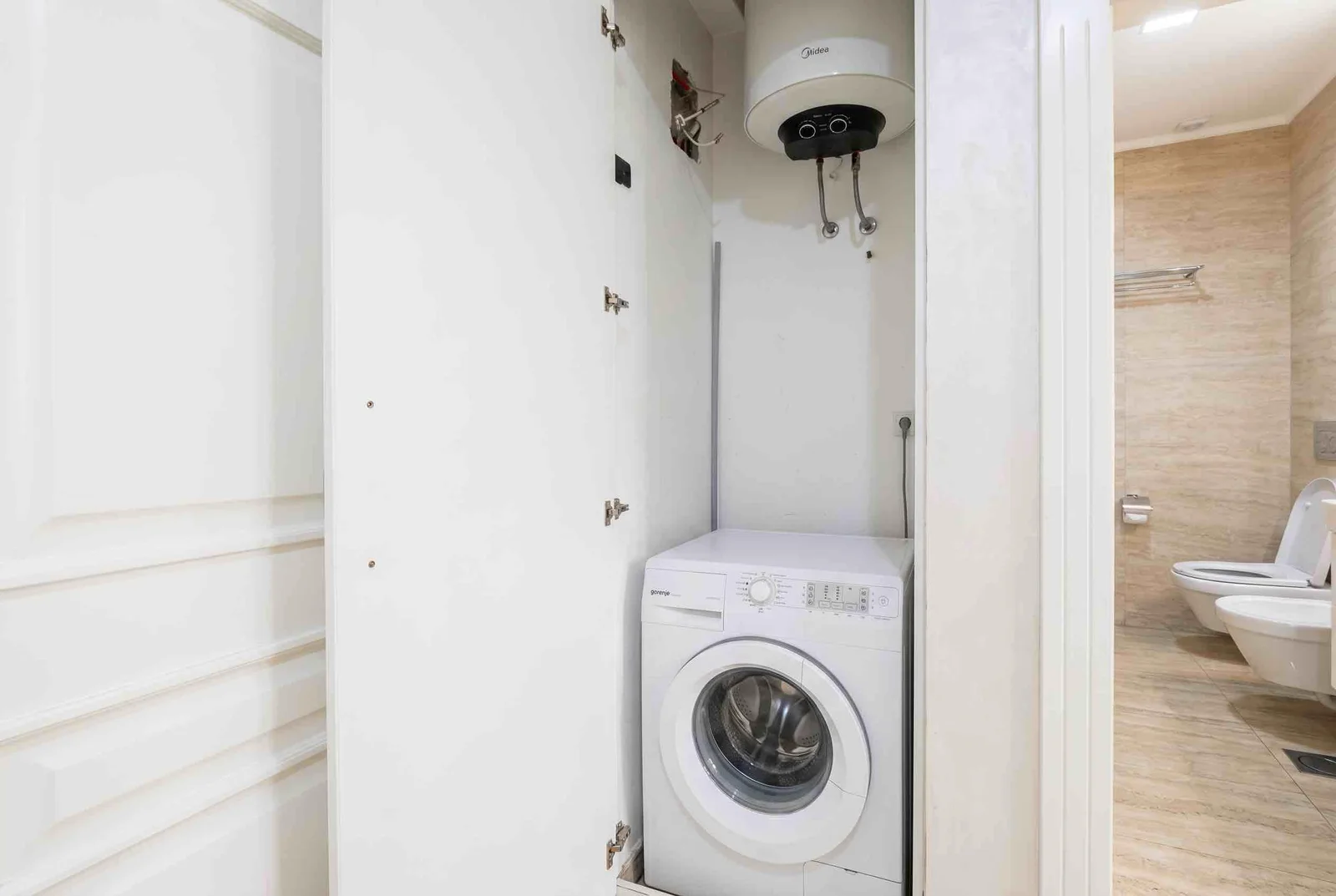 Washing machine in a niche with a water heater and a visible toilet in the adjacent room.
