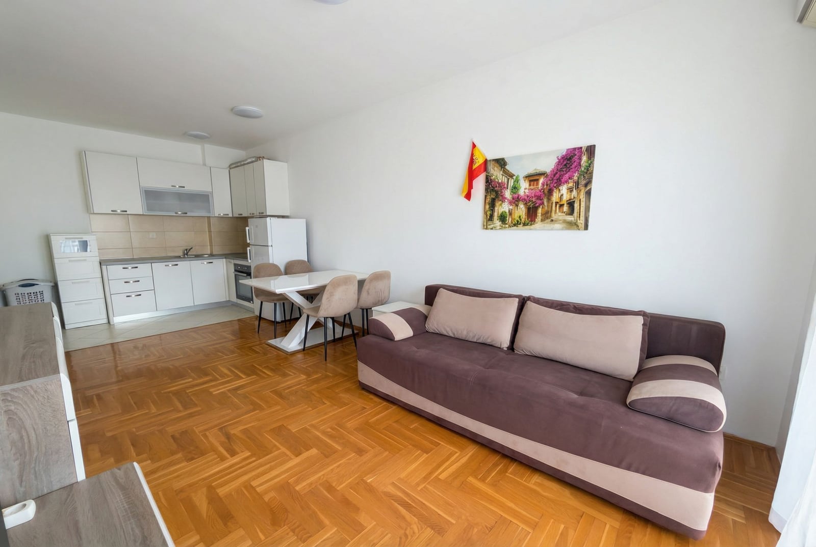 Dnevna soba sa kuhinjom u stanu u Budvi, Crna Gora: braon sofa, trpezarijski deo i svetao kuhinjski nameštaj.