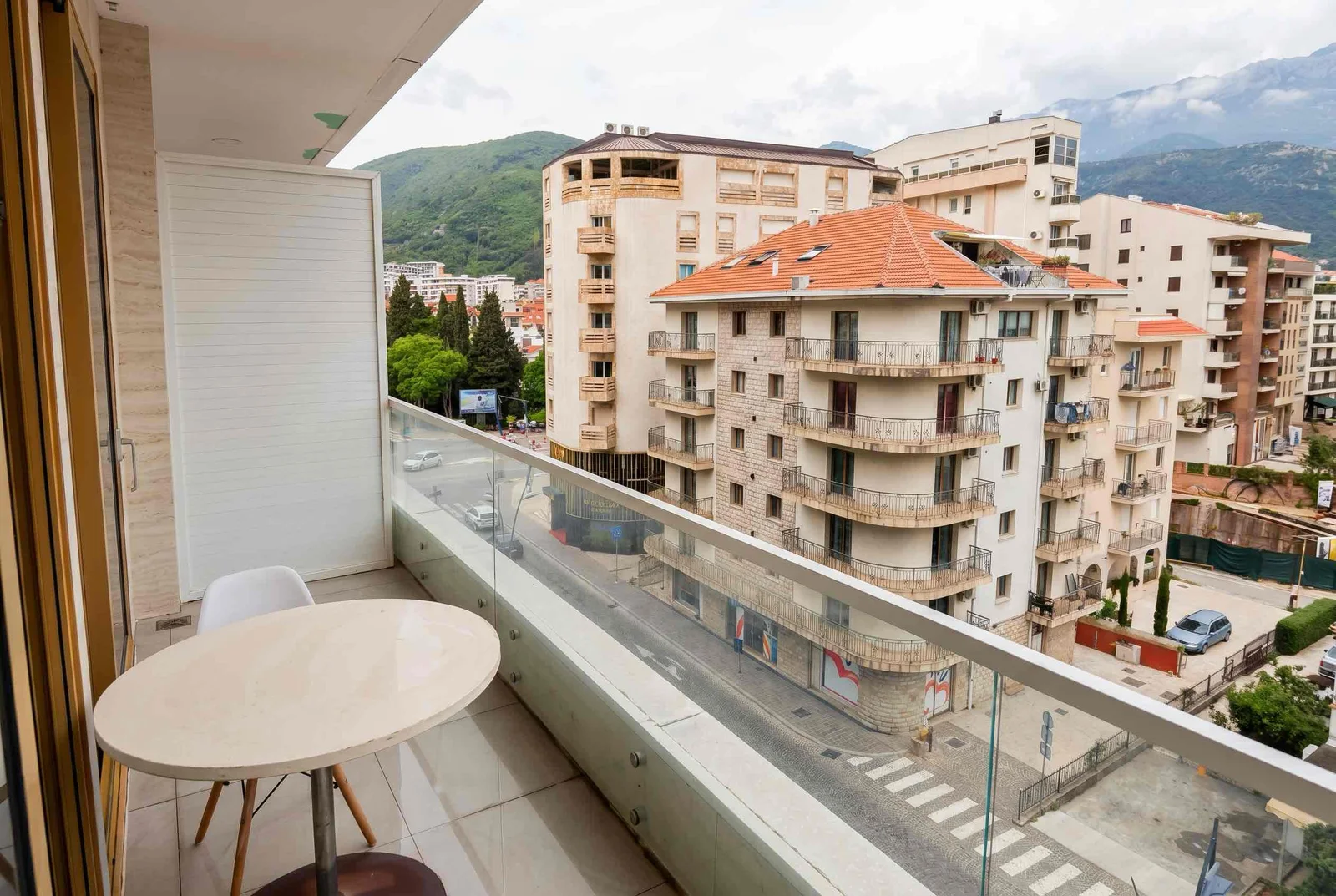 Balcony with a white table and glass railing overlooking city buildings and mountains.