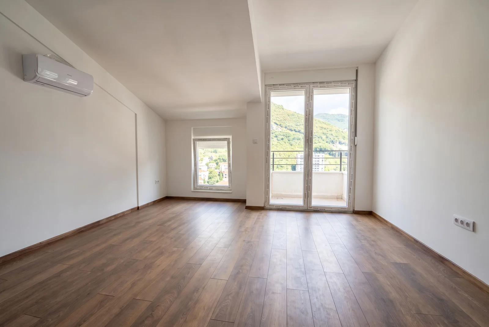 Pusta soba sa drvenim podom, klimom i vratima na balkon sa vidikom na brda.