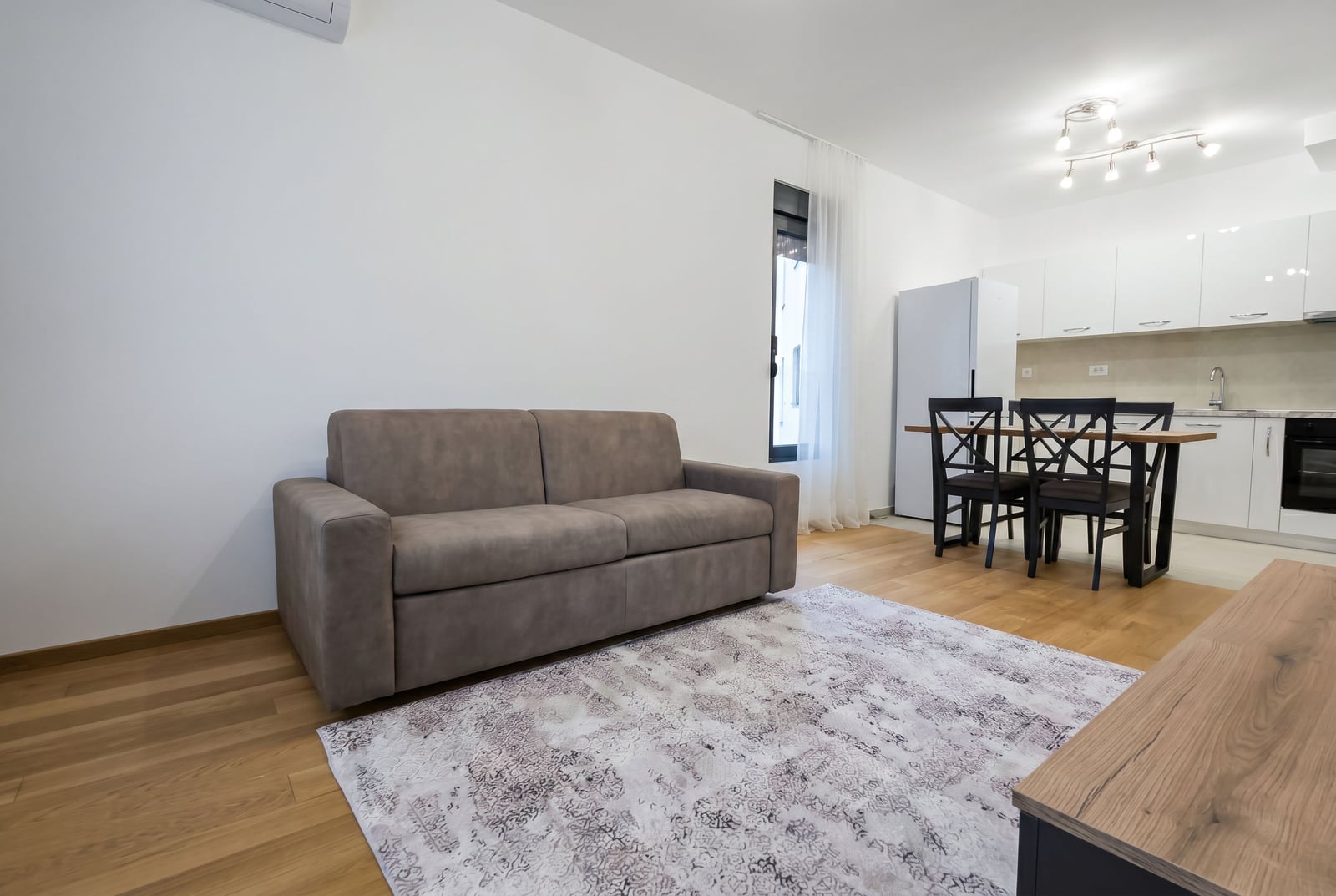 Modern living room with a sofa and kitchen in an apartment in Bečići, Montenegro: bright interior, parquet flooring, and dining area.