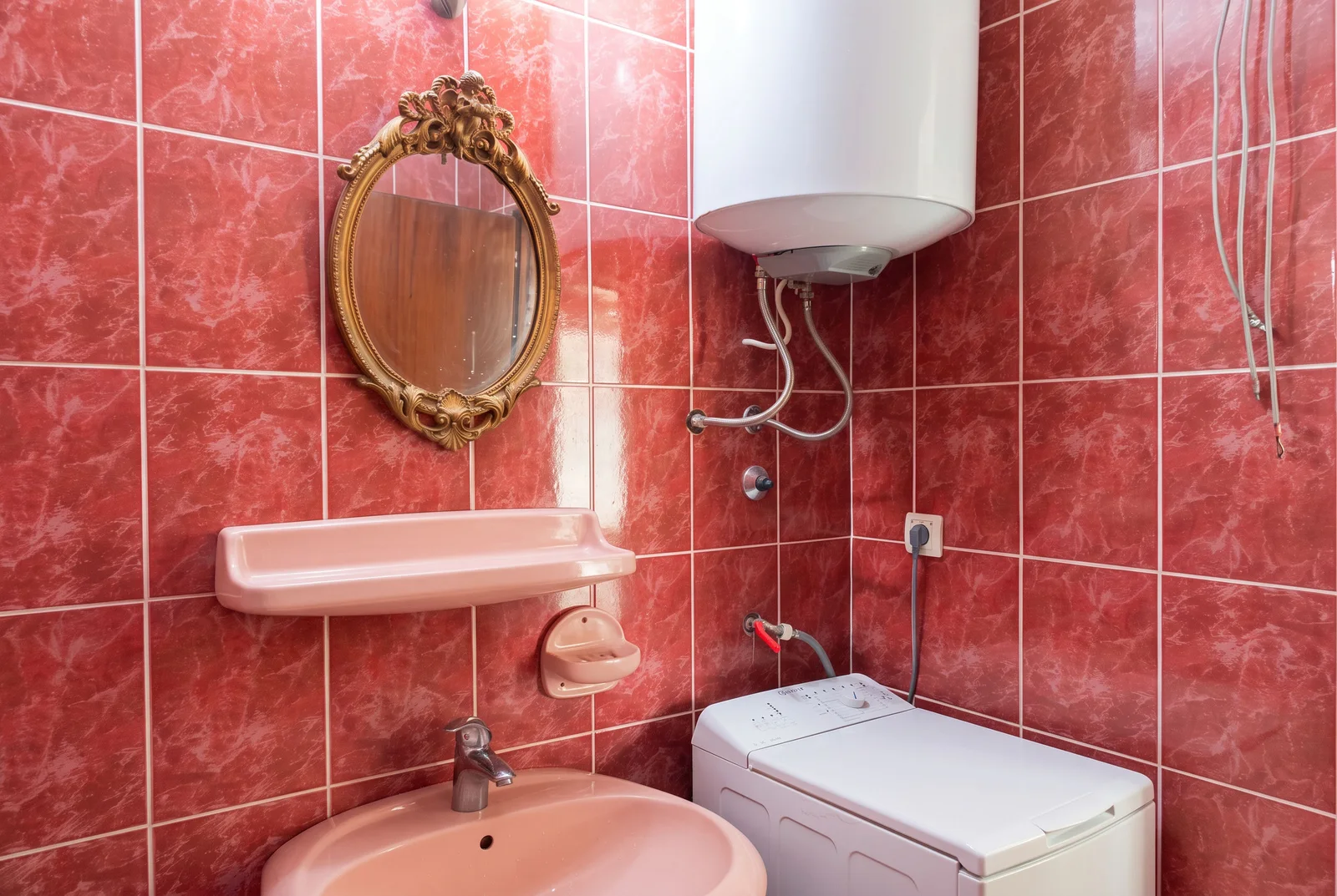 Red tiles, pink sink, gold mirror, wall-mounted water heater, and a white washing machine in the corner.