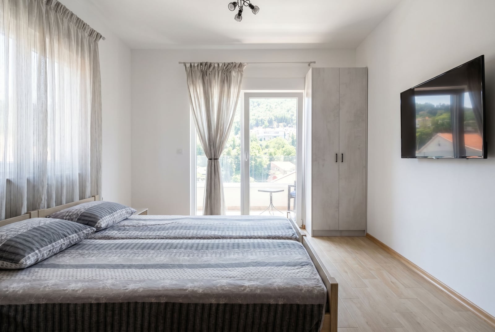Svetla spavaća soba u stanu u Budvi, Crna Gora: bračni krevet, ormar, TV i izlaz na balkon sa pogledom na planine.