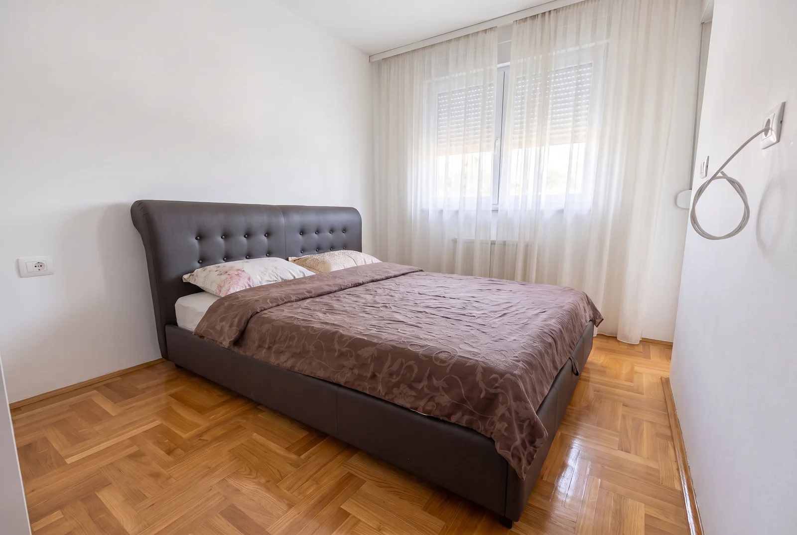 Spavaća soba sa dvosmernim krevetom, sivim naslonom, drvenim podom i belim zavesama na prozoru.