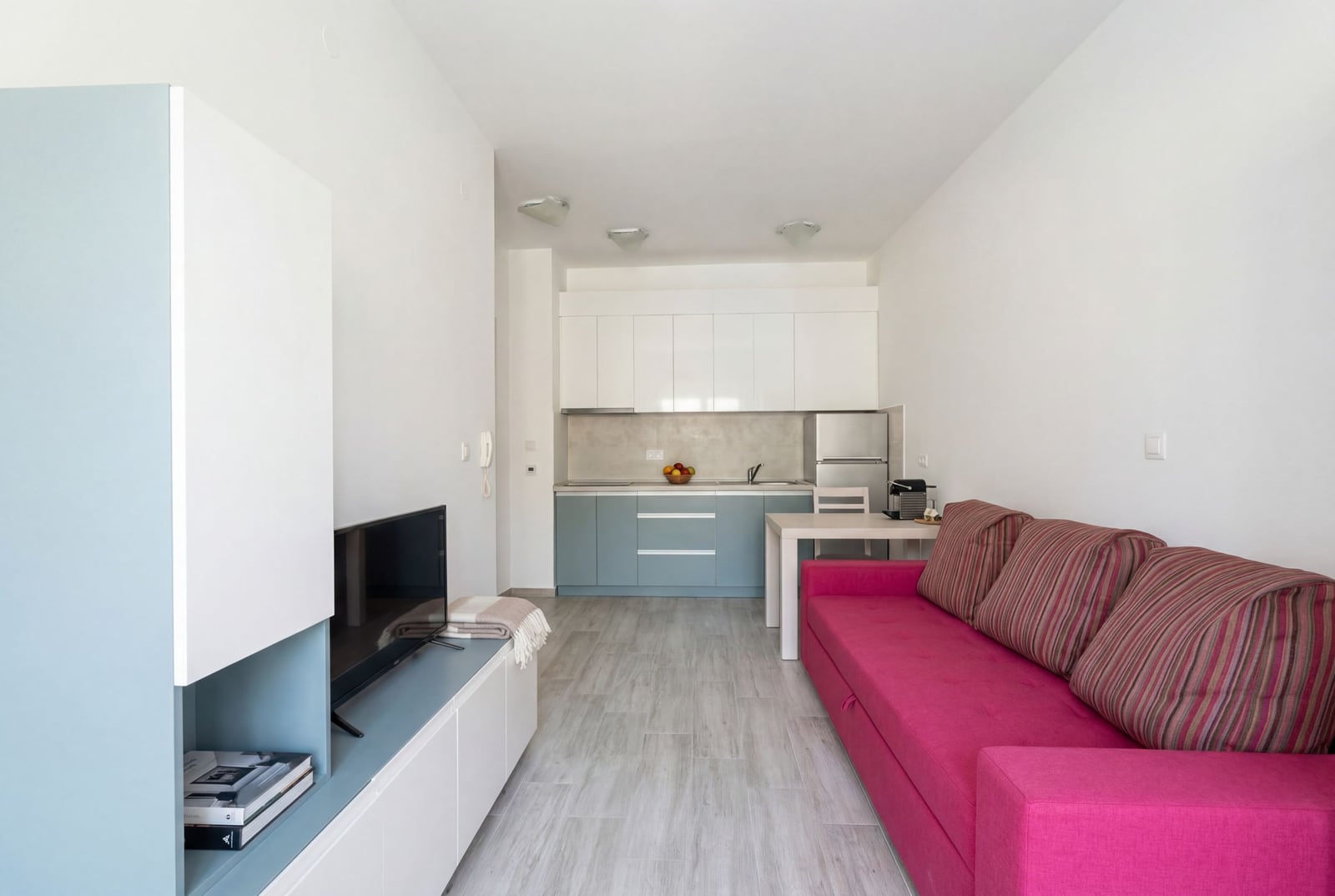 Modern living room with kitchen in an apartment in Budva, Montenegro: bright pink sofa, white furniture, and dining area.