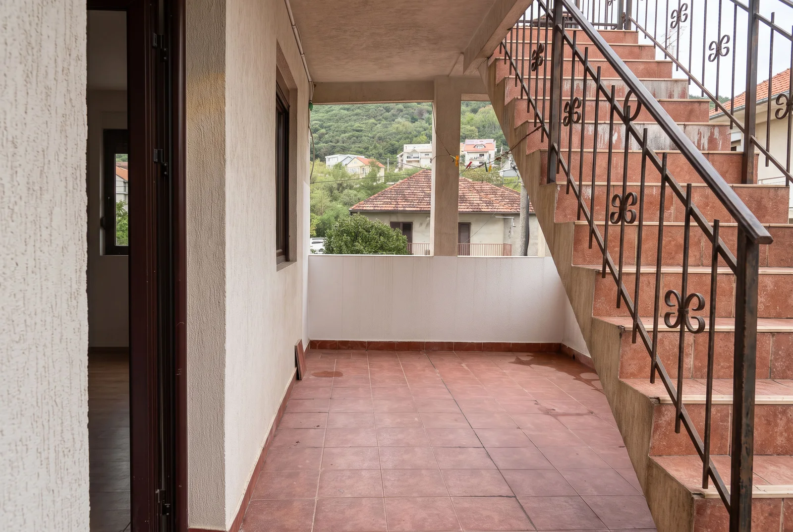 Terrace with red tiles and a staircase featuring wrought iron railings, overlooking houses and greenery.