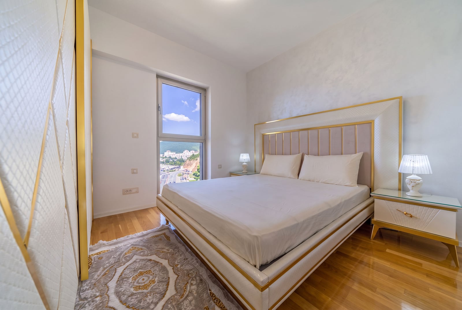 Bright bedroom with a double bed, nightstands, and a view of the city in Bečići, Montenegro.