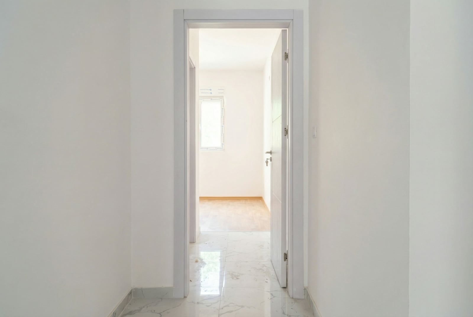 Svetao hodnik sa belom završnom obradom i pločicama na podu u stanu u Budvi, Crna Gora, pogled na praznu sobu sa prozorom.
