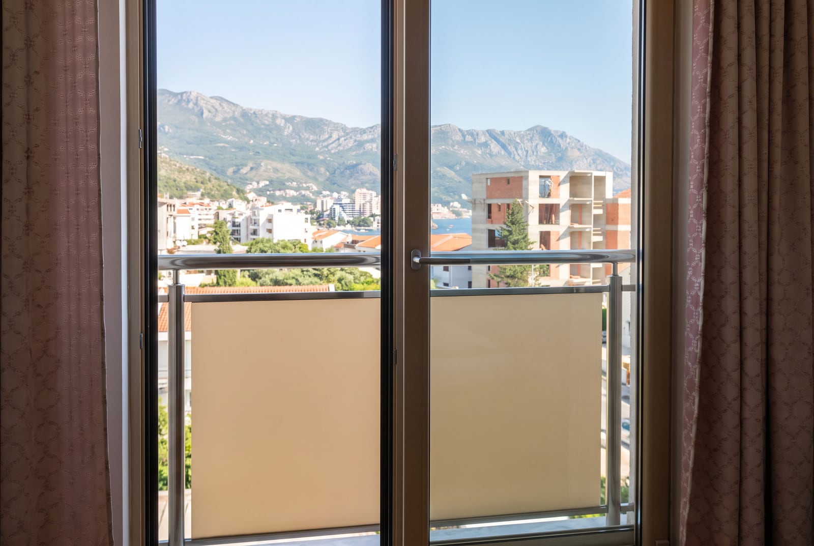 View from the bedroom window of an apartment in Bečići, Montenegro: a panorama of mountains, city buildings, and the sea.