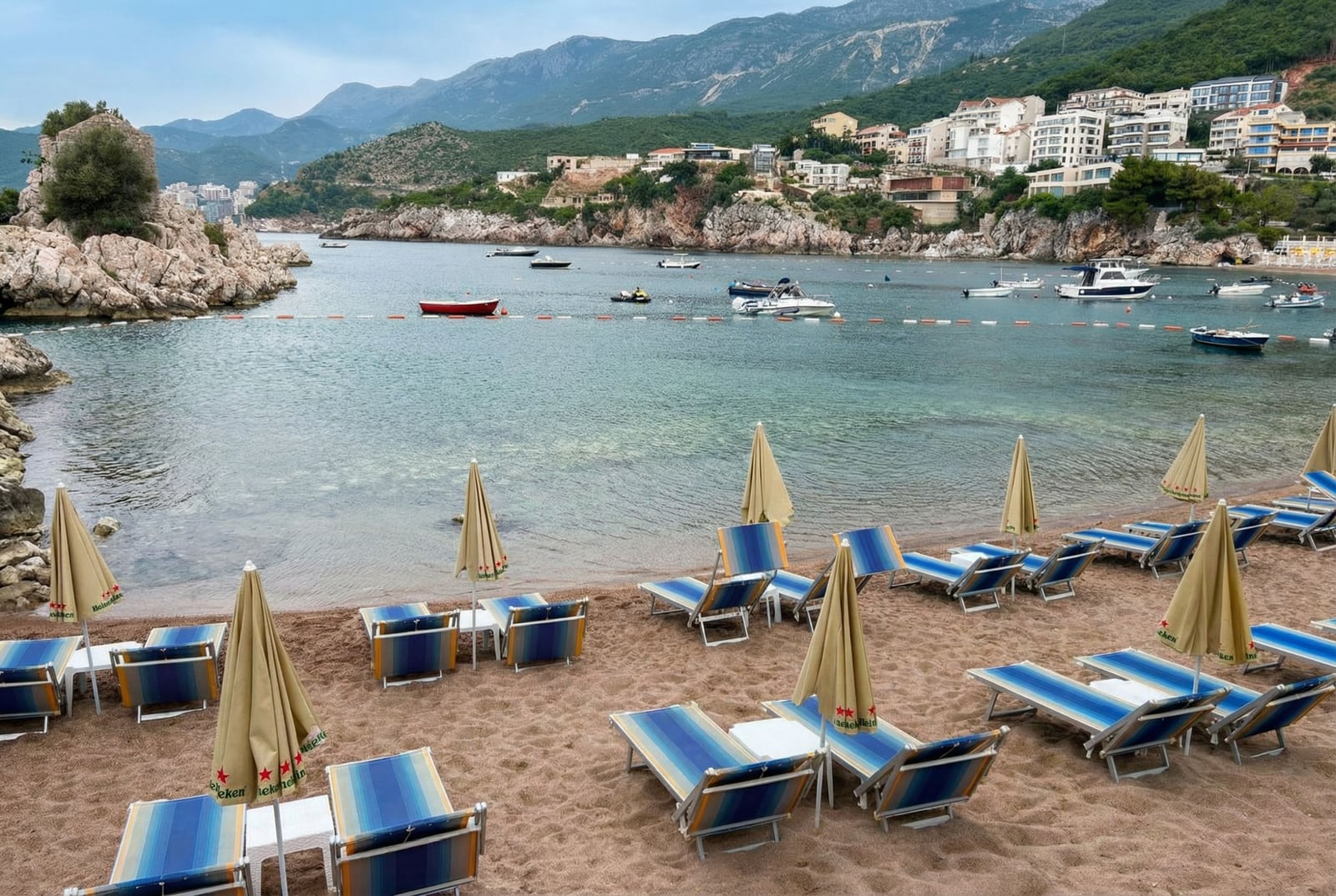 Peščana plaža sa ležaljkama i suncobranima u Pržnu, Crna Gora, s pogledom na slikovitu uvalu, stene i usidrene čamce.