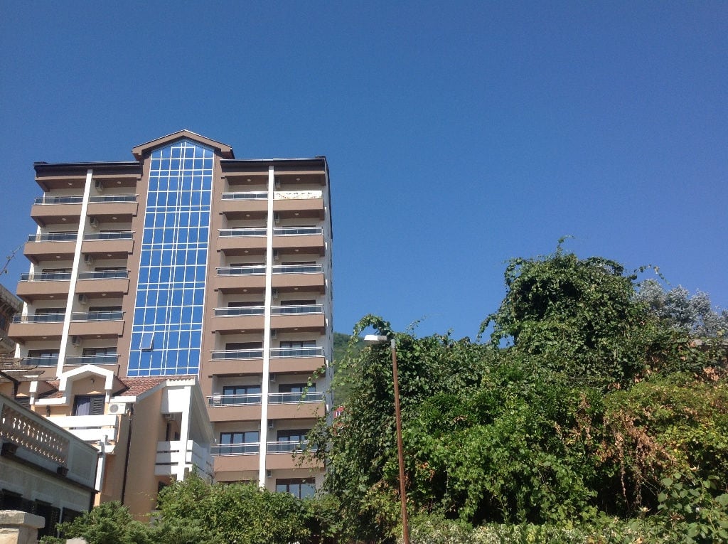 Modern multi-story residential building with panoramic glazing in Budva, Montenegro, against a clear blue sky.