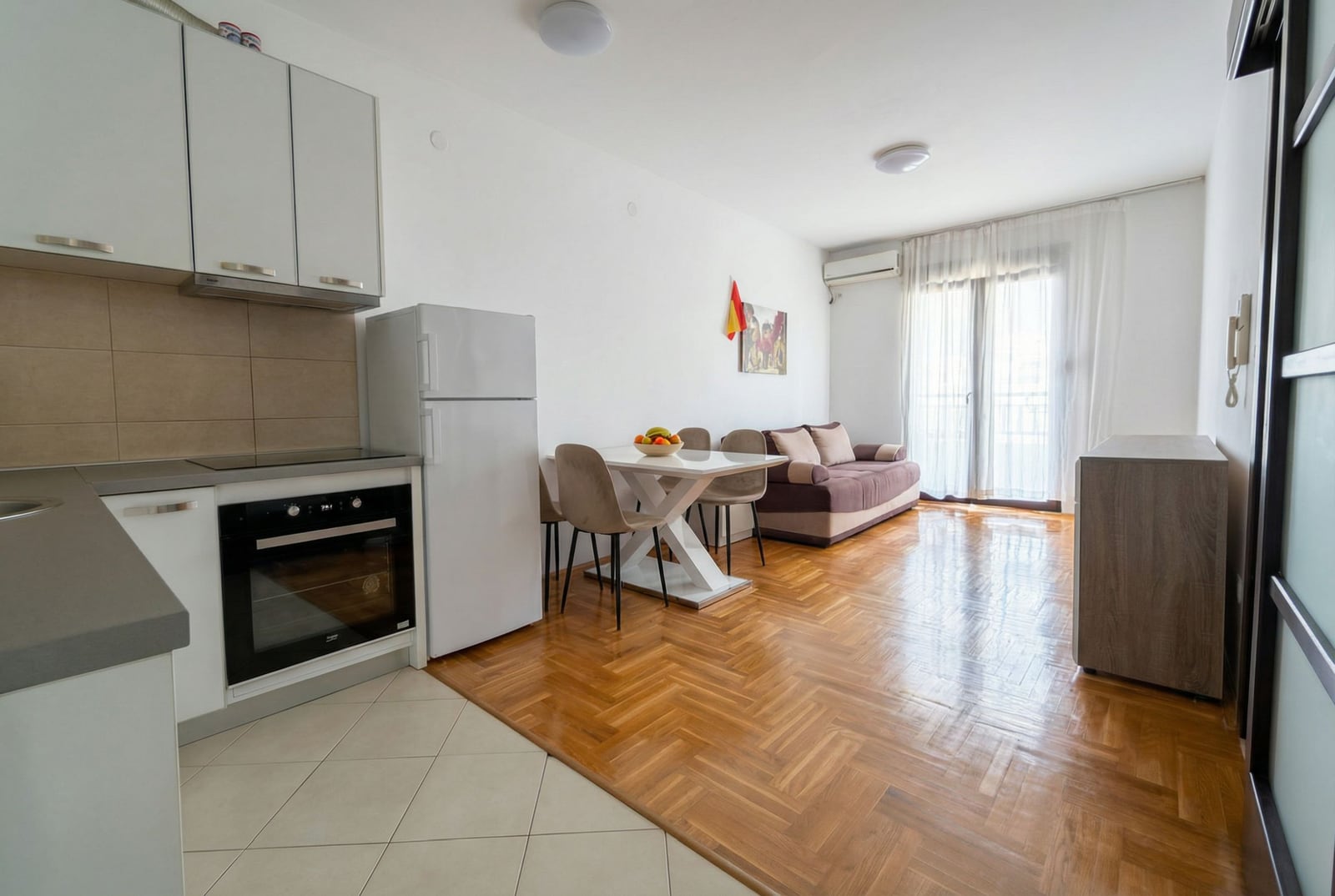 Savremeni dnevni boravak sa kuhinjom u stanu u Budvi, Crna Gora: svetao nameštaj, parket i izlaz na balkon.