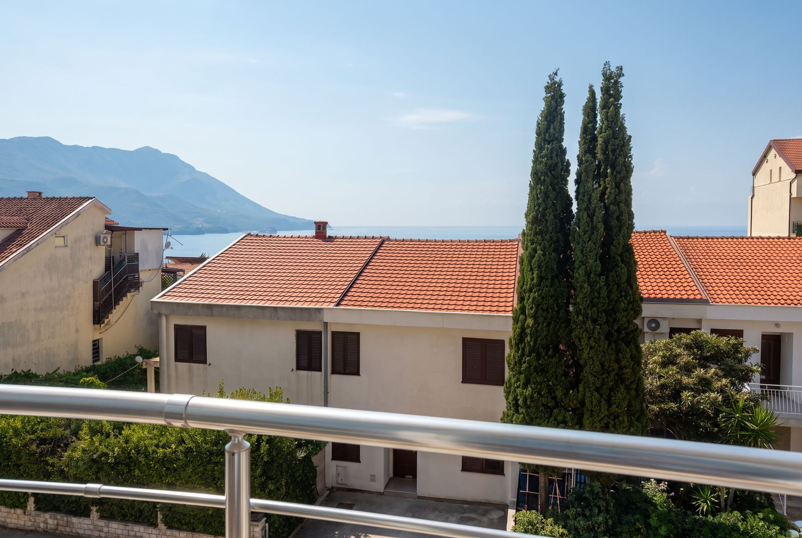 Pogled sa balkona stana u Bečićima, Crna Gora: krovovi od crepa, zeleni čempresi, planine i more.