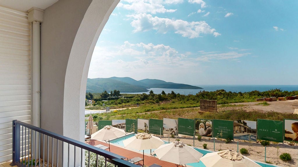 Pogled sa balkona stana u Luštici, Crna Gora: bazen sa suncobranima, panorama planina i Jadranskog mora.