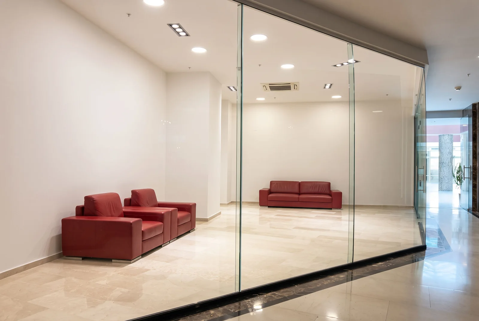 Modern lobby or office space featuring red leather sofas, glass partition walls, and polished marble flooring under recessed lighting.
