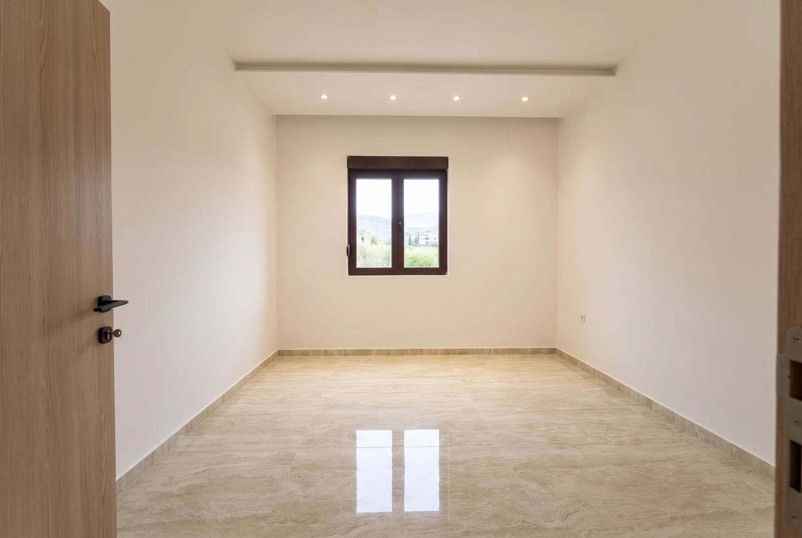 Empty room with beige tiled floors, white walls, recessed ceiling lights, and a window overlooking a natural landscape.